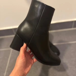 Svarta stövletter med blockklack - Snygga svarta stövletter i skinnimitation med blockklack på ca 7 cm. Klassisk och stilren design med rund tå och dragkedja på sidan. Perfekta till både jeans och klänning för en trendig look. 