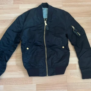 Svart bomberjacka Alpha Industries - Svart bomberjacka från Alpha Industries med klassisk guldfärgad dragkedja och ribbade muddar. Jackan har en snygg detalj med 'Remove Before Flight'-tag på ärmfickan och två fickor med guldfärgade knappar. Tillverkad i slitstark polyester, perfekt för dig som gillar streetstyle.