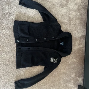 Svart kofta från Polo Ralph Lauren - Svart stickad kofta från Polo Ralph Lauren med coolt dödskallemärke på bröstet. Koftan har knappar framtill, ribbade muddar och en klassisk krage. 