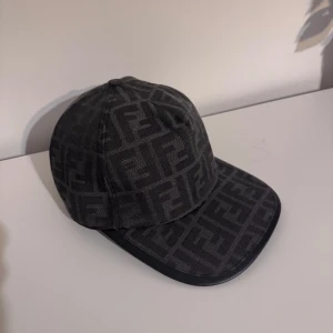 Svart keps från Fendi med FF-mönster - Snygg svart keps från Fendi med det ikoniska FF-logomönstret över hela kepsen. Klassisk böjd skärm med svart kant och justerbar rem baktill där Fendi Roma är broderat. Perfekt accessoar för att lyfta din streetwear-look. Priset är ej hugget i en sten då kepsen är helt oanvänd.
