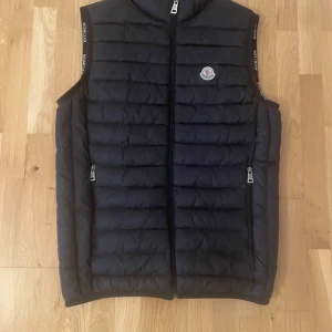 Svart dunväst från Moncler - Snygg svart dunväst från Moncler med quiltad design och dragkedja framtill. Västen har två fickor med dragkedja och ikonisk Moncler-logga på bröstet. Insidan är fodrad i rött med coolt serietryck. Perfekt för lager-på-lager och streetstyle.