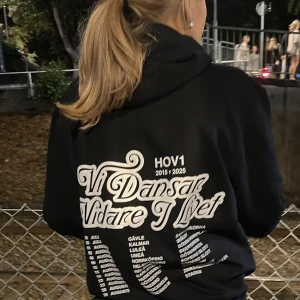 Svart Hov1 hoodie med tryck - Svart hoodie från Hov1 med vit kronlogga framtill och stort turnétryck på ryggen med texten 'Vi Dansar Vidare I Livet' och turnédatum. Klassisk känguruficka och snörning i huvan. Perfekt för dig som älskar svensk musik och streetstyle.