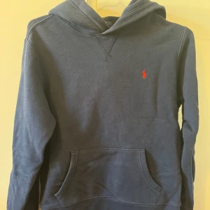 Mörkblå hoodie från Polo Ralph Lauren - Klassisk mörkblå hoodie från Polo Ralph Lauren med röd broderad logga på bröstet. Barnstorlek. Tröjan har huva, känguruficka och är tillverkad i mjuk bomullsmix. Perfekt för en chill och stilren look. 