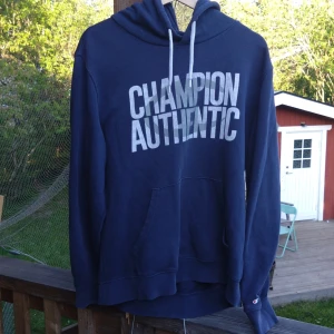 Mörkblå hoodie från Champion - Säljer en mörkblå hoodie från Champion med texten 'CHAMPION AUTHENTIC' tryckt i vitt på bröstet. Tröjan har huva med dragsko och långa ärmar. Perfekt för en avslappnad stil. Kontakta mig gärna ifall du har några funderingar, pris kan diskuteras. 