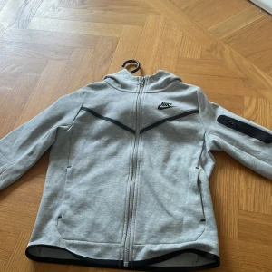 Grå zip-hoodie från Nike - Säljer en grå hoodie från Nike med hel dragkedja och svart Nike-logga på bröstet. Jackan har huva, svarta detaljer och en ficka med dragkedja på ena ärmen. Perfekt för en sportig look.priset är inte fast hör av er vid frågor eller undringar 