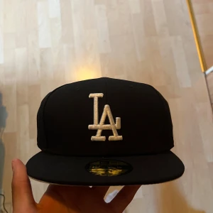 Svart LA-keps från New Era - Säljer en svart keps från New Era med LA-logga broderad i vitt framtill. Klassisk modell med platt skärm och broderad MLB-logga bak. Perfekt för dig som gillar sportig stil och streetwear. Säljer då den ej passade