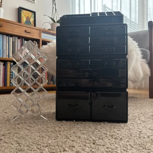 Sminkställ - Säljer en svart sminkförvaring i plast med flera små och stora lådor, perfekt för att organisera smink som läppstift, borstar och ögonskuggor. Medföljer även ett transparent ställ för läppstift eller läppglans.