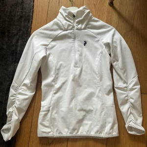 Vit långärmad träningströja med dragkedja - Säljer en vit långärmad träningströja från Peak Performance. Tröjan har halv dragkedja framtill, hög krage och rynkade detaljer längs ärmarna. Perfekt för träning eller som ett extra lager under kyliga dagar.