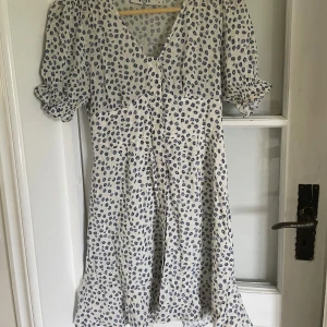 Vit och blå mönstrad klänning från NA-KD - Säljer en söt vit klänning med blått blommigt mönster från NA-KD i storlek 34. Klänningen har korta puffärmar, v-ringning och knappar framtill.