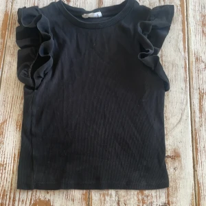 Svart ribbad topp med volangärmar från Terranova - Säljer en svart ribbad topp från Terranova med volangdetaljer på axlarna. Toppen är ärmlös och har en rund halsringning. Perfekt för att lyfta en enkel outfit med lite extra stil.