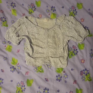 Ljusblå blommig blus med rynkade detaljer - Supersöt ljusblå blus med små vita blommor och rynkade detaljer framtill. Blusen har korta puffärmar och smockad rygg för extra komfort. Perfekt för vår och sommar!