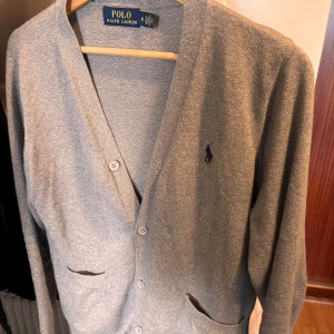 Grå kofta från Polo Ralph Lauren - Säljer en klassisk grå kofta från Polo Ralph Lauren med knäppning framtill och två fickor. Tröjan har lång ärm och den ikoniska broderade loggan på bröstet. Perfekt för lager-på-lager och stilrena outfits.