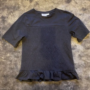 Marinblå topp med tightningar  - Marinblå topp från Gina Tricot med korta ärmar och smockad struktur på framsidan och baksidan. Nedtill finns en volangkant som ger en söt touch. Tillverkad i mjuk bomull, perfekt för en trendig och bekväm stil. Använd 2 gånger.