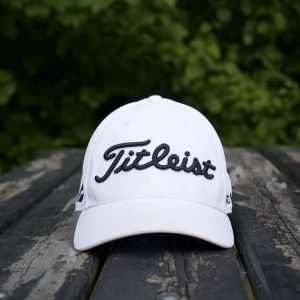 Vit keps från Titleist - Snygg vit keps från Titleist med svart broderad logga framtill och detaljer på sidorna, inklusive 'PRO V1' och 'FJ'. Kepsen har böjd skärm och klassisk passform, perfekt för golf eller vardag.