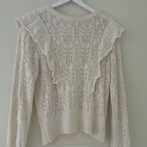 Beige stickad tröja med volang från Ellos - Supersöt beige stickad tröja från Ellos med volangdetaljer framtill och ett fint hålmönster. Tröjan har långa ärmar och bred ribbad kant nedtill och vid ärmsluten. Perfekt för lager-på-lager och en mysig look. Jättebra skick! Köpt för 400.