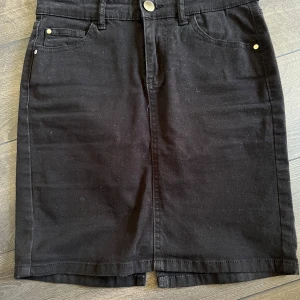 Svart jeanskjol från Cubus - Svart jeanskjol från Cubus i klassisk femficksmodell med knapp och dragkedja framtill. Kjolen är rak i modellen och har slits bak för extra rörelsefrihet. Tillverkad i bomull med lite stretch för skön passform.