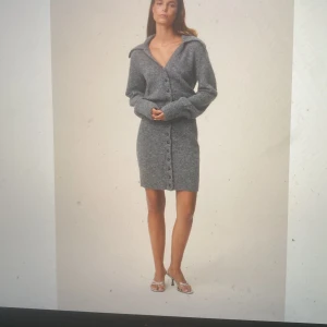 Grå stickad klänning med knappar från Adoore - Superfin grå stickad klänning från Adoore med långa ärmar och stora krage. Klänningen har knappar hela vägen framtill och en figurnära passform som markerar midjan. Perfekt för dig som gillar en stilren och mysig look.