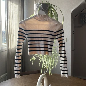 Randig croppad tröja från Pull&Bear - Säljer en croppad ribbad tröja från Pull&Bear i vitt med svarta horisontella ränder. Tröjan har lång ärm och hög hals, perfekt för en trendig vardagslook.