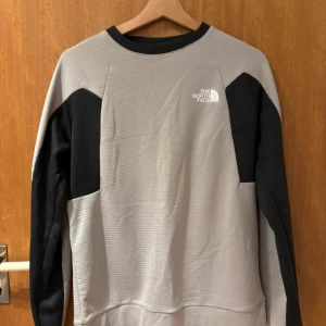 Grå och svart tröja från The North Face - Säljer en stilren grå tröja med svarta detaljer från The North Face. Tröjan har rund halsringning och långa ärmar. Perfekt för träning eller vardag. Logga på bröstet och ärmen.