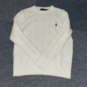Vit kabelstickad tröja från Polo Ralph Lauren - Vit kabelstickad tröja från Polo Ralph Lauren med klassisk broderad logga på bröstet. Tröjan har rund halsringning och ribbade muddar vid ärmslut och nederkant. Perfekt för lager-på-lager under kyliga dagar.