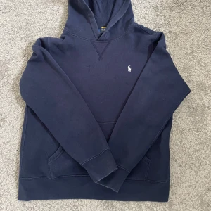 Mörkblå hoodie från Polo Ralph Lauren - Mörkblå hoodie från Polo Ralph Lauren med klassisk huva och magficka. Tröjan har en liten vit broderad logga på bröstet och är tillverkad i mjukt material. Perfekt för en avslappnad stil.