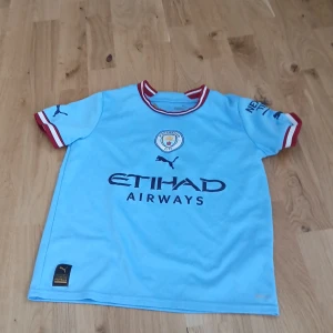 Manchester City fotbollströja Haaland 9 - Ljusblå Manchester City fotbollströja från Puma med Haaland 9 på ryggen. Tröjan har korta ärmar med röda och vita detaljer vid ärmslut och krage. Klubbmärke och sponsorlogga på bröstet. Tillverkad i lätt och ventilerande material.