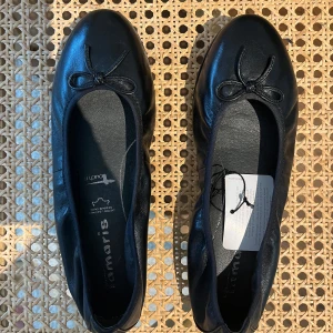 Svarta ballerinaskor Tamaris - Säljer ett par klassiska svarta ballerinaskor från Tamaris i läder med rund tå och en liten rosett framtill. Skorna har platt sula och är perfekta för sommaren! 😋🫧🌷Har inte använts och därmed nyskick