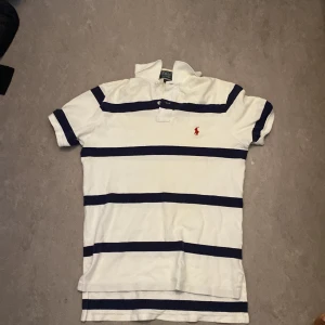 Polo Ralph Lauren Vit och marinblå randig pikétröja  - Vit pikétröja med breda marinblå ränder från Polo Ralph Lauren. Klassisk krage och knappslå framtill, samt röd broderad logga på bröstet. Modellen är custom fit och har korta ärmar. Perfekt för en sportig och stilren look.