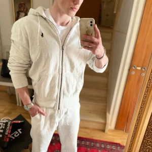 Vit hoodie från Polo Ralph Lauren - Säljer en stilren vit hoodie från Polo Ralph Lauren med dragkedja och huva. Jackan har ett litet broderat logomärke på bröstet och dragsko vid huvan. Perfekt för en avslappnad och sportig look.