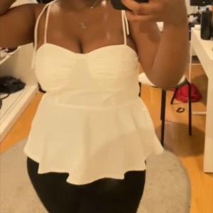 Vit peplumtop med tunna axelband - Säljer en söt vit peplumtop med tunna knytbara axelband och markerad byst. Toppen har en lätt utsvängd nederdel och är perfekt för varma dagar.