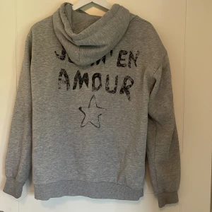 Grå hoodie med tryck 'JE M'EN AMOUR' - Säljer en grå hoodie med huva och tryck både fram och bak där det står 'JE M'EN AMOUR' samt en stjärna. Tröjan har ribbade muddar och är i mjukt material, perfekt för en avslappnad stil.