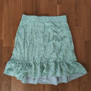 Grön blommig kjol med volang - Söt ljusgrön kjol med småblommigt mönster och volang nertill. Kjolen är något längre bak än fram, vilket ger en fin siluett. Perfekt för vår och sommar!