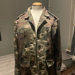 Camouflage vindjacka med dragkedja och knappar - Säljer en militärinspirerad camouflage vindjacka med både dragkedja och knappar framtill. Jackan har flera fickor, axelklaffar och klassisk krage. Perfekt för dig som gillar streetwear eller vill ha en cool lager-på-lager-look.