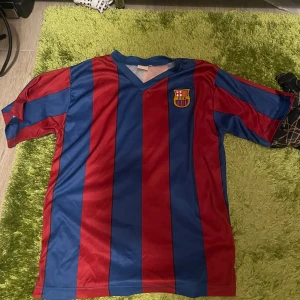 FC Barcelona fotbollströja Ibrahimović 9 - Säljer en klassisk FC Barcelona fotbollströja med röda och blå breda ränder, klubbmärke på bröstet och Ibrahimović 9 tryckt i gult på ryggen. Tröjan har V-ringning och är kortärmad. Perfekt för fans av Zlatan och Barca!