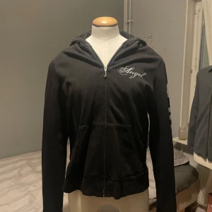 Svart hoodie från Juicy Couture med textdetaljer - Svart hoodie med dragkedja från Juicy Couture. På bröstet står det 'Angel' i vitt och på ärmen finns stora vita tryck med hjärtan och texten 'JUICY'. Tröjan har huva och fickor framtill.