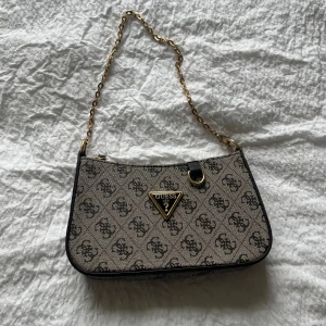 Axelväska från Guess - Säljer en beige och svart axelväska från Guess med kedjerem och logomönster över hela väskan. Väskan har guldfärgade detaljer och ett utbytbart axelband som är justerbart i svart. 