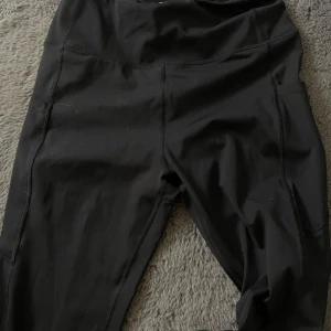 Svarta träningstights från Energetics - Svarta tights från Energetics med hög midja och praktiska sidofickor. Perfekta för träning med stretchigt material som ger bra rörelsefrihet. Snygga sömdetaljer längs benen och diskret logga på höften. Står storlek M men sitter bra på storlek S.