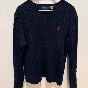 Mörkblå kabelstickad tröja från Polo Ralph Lauren - Säljer en klassisk mörkblå kabelstickad tröja från Polo Ralph Lauren med rund halsringning och röd broderad logga på bröstet. Jag säljer för att jag knappt använder längre. Hör av er vid frågor