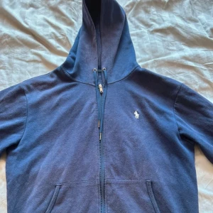 Marinblå hoodie från Polo Ralph Lauren - Säljer en marinblå hoodie från Polo Ralph Lauren med dragkedja och klassisk broderad logga på bröstet. Tröjan har huva, fickor framtill och långa ärmar. Perfekt för en avslappnad stil.