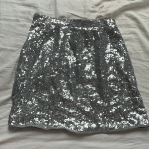 Silver paljettkjol från Gina Tricot - Säljer en glittrig silverfärgad kjol från Gina Tricot, täckt med paljetter för en riktigt festlig look. Kjolen är kort och har en rak passform som sitter snyggt i midjan. Perfekt för dig som vill sticka ut! Paljetter har gått av lite på olika ställen men syns inte på långt håll!