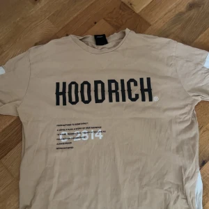 Beige t-shirt från Hoodrich - Säljer en beige t-shirt från Hoodrich med stor svart logga på bröstet och vit textdetalj nedtill. Klassisk rund hals och korta ärmar. Perfekt för en avslappnad streetwear-look.