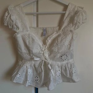 Vit blommig blus med volang och knyt - Superfin vit blus med broderade blommor, volangkant och knyt framtill. Puffiga korta ärmar och v-ringad halsringning. 