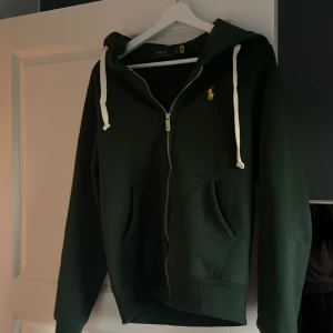 Mörkgrön hoodie från Polo Ralph Lauren - Säljer en mörkgrön hoodie från Polo Ralph Lauren med dragkedja och gul broderad logga på bröstet. Tröjan har huva med vita snören och två fickor framtill. Perfekt för en avslappnad stil. Har använt den få gånger passar bra storlek xs/s