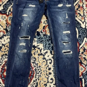 Dsquared2 Blå jeans, Storlek 44, PRISET KAN DISKUTERAS - Säljer ett par blå jeans med flera slitningar och hål framtill samt vita färgstänk för en riktigt edgy look. Jeansen har rak passform och klassisk femficksdesign. Perfekta för dig som gillar streetstyle.