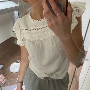 Vit blus med spetsdetaljer - Säljer en söt vit blus med volangärmar och vackra spetsdetaljer vid halsringningen och axlarna. Blusen har en lös passform och knytning nertill. Perfekt för vår och sommar!