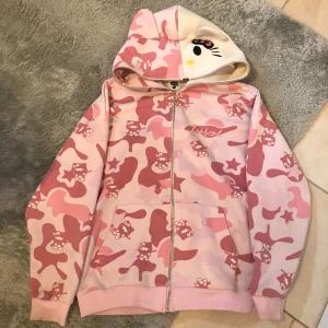 Rosa camo hoodie med Hello Kitty-öron - Säljer en söt hoodie med rosa och ljusrosa camouflage-mönster och Hello Kitty-motiv. Huvan har öron och broderat ansikte. Dragkedja framtill och två fickor. Perfekt för dig som gillar gulliga och unika plagg!