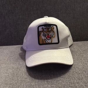 Vit keps med tigerlogga - Vit keps med böjd skärm och SnapBack baktill. Framtill finns en broderad patch med en tiger och texten 'TIGER'. Justerbar passform med plastspänne bak. Perfekt för en cool och sportig look.
