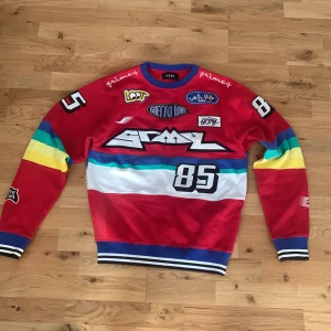 Färgglad sweatshirt från GRMY med tryck - Säljer en röd sweatshirt från GRMY med färgglada ränder på ärmarna och bröstet. Tröjan har flera tryck och patches, bland annat siffran 85 och olika loggor. Ribbstickade muddar i blått och vitt vid ärmslut och nederkant.