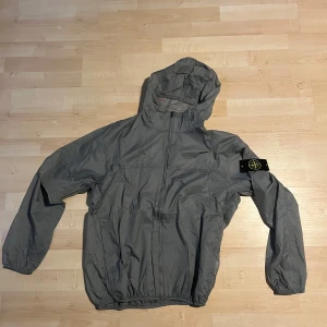 Grå vindjacka från Stone Island - Säljer en grå vindjacka från Stone Island med huva och dragkedja framtill. Jackan har den klassiska logotypen på vänster ärm och elastiska muddar vid ärmslut och midja. Perfekt för blåsiga dagar. Oanvänd, pris kan diskuteras