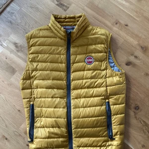 Gul dunväst från Colmar - Snygg gul dunväst från Colmar med svart dragkedja och två fickor framtill. Västen är i storlek 50 men de motsvarar storlek M tror jag!! Nästan aldrig använd!!!